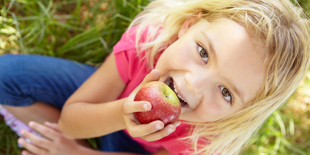 Mädchen sitzt im Grünen und beißt mit gesunden Zähnen in einen Apfel